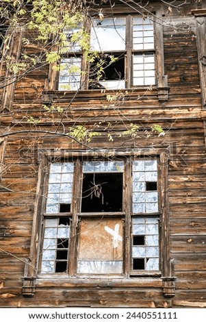 The wooden house is in disrepair with broken windows and boarded-up doors. The paint is peeling, and the wood is rotting. The house is surrounded by overgrown plants and trees. Royalty-Free Stock Photo #2440551111