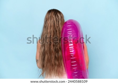 Young blonde woman in swimsuit holding inflatable donut isolated on blue background in back position