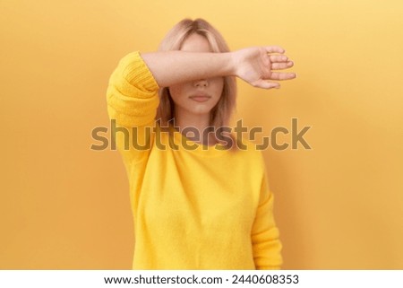 Young caucasian woman wearing yellow sweater covering eyes with arm, looking serious and sad. sightless, hiding and rejection concept  Royalty-Free Stock Photo #2440608353
