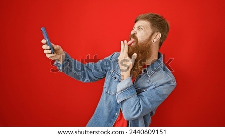 Cool irish guy with a beard, young redhead adult making a selfie over isolated red background using smartphone technology, casual lifestyle expression standing alone, looking handsome.
