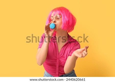 Beautiful young woman in pink wig with blue sweet macaroon showing heart gesture on yellow background Royalty-Free Stock Photo #2440620661