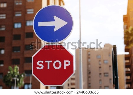 Traffic rules. Red road sign stop and blue and white right directional arrow on residential buildings background. Street sign direction pointing turn right, but need to stop before. Two road signs Royalty-Free Stock Photo #2440633665