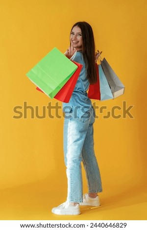 Standing and holding shopping bags. Young woman is against yellow background.