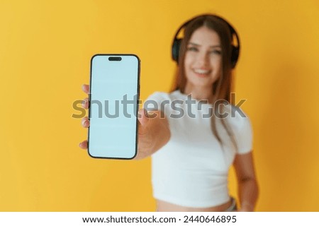 Headphones on head, holding smartphone with copy space. Young woman is against yellow background.