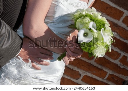 The image beautifully frames the clasped hands of a bride and groom against the backdrop of her elegant bridal bouquet and the timeless texture of a brick wall, capturing a moment of unity and Royalty-Free Stock Photo #2440654163