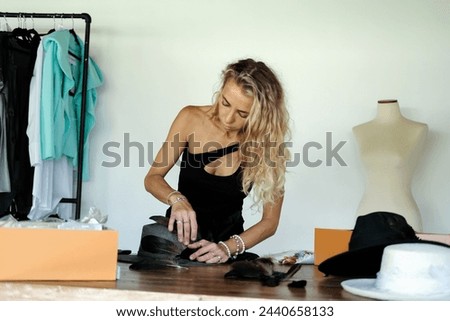 A woman hatmaker in a workshop decorates a hat with feathers and threads. Headwear designer at work. Fashion designer's new hat collection. Royalty-Free Stock Photo #2440658133