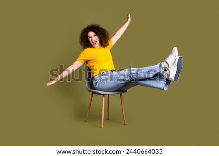 Full length photo of young overjoyed woman in yellow t shirt and jeans flying like airplane sit chair isolated on khaki color background