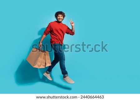 Full length photo of young mexican guy jumping v sign symbol with packages greetings at shopping mall isolated on blue color background