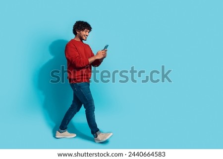 Full body length photo of funny mexican guy in red sweatshirt walking with smartphone texting someone isolated on cyan color background