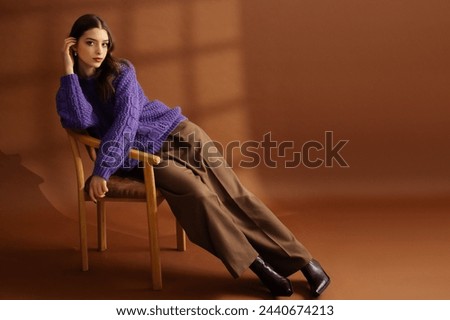 Fashionable confident beautiful brunette woman wearing trendy purple knitted sweater, pants, leather boots, posing on brown background. Studio fashion portrait. Copy, empty, blank space for text