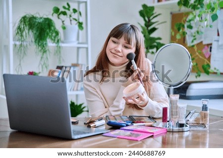 Young female showing cosmetics to laptop webcam, sitting at table at home