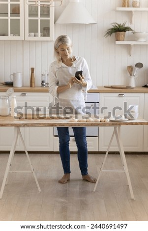 Smiling middle aged housewife stand at kitchen table with smartphone, check online recipe, take picture of baking process on mobile phone, chatting, enjoy cooking time at home. Modern tech, culinary