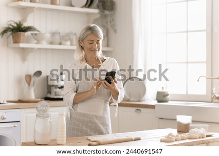 Smiling middle aged housewife stand at kitchen table with smartphone, check on-line recipe, take picture of baking process on mobile phone, chatting, enjoy cooking time at home. Modern tech, culinary