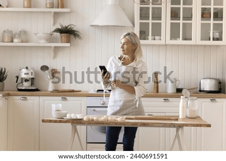 Pretty middle-aged woman using cellphone standing in modern kitchen, distracted from cooking, wait for call, answers on sms, chatting, send message, search recipes through on-line cookbook application