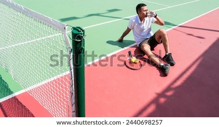 Young latín man tennis player resting and drinking water on tennis court in sunny hot day at summer