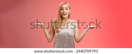 Carefree fascinated young blond girl 20s in silver glittering evening party dress raising hands sideways weighing shrugging smiling amused search right choice, deciding what do red background.