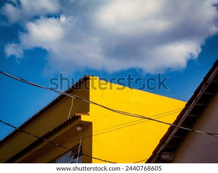 An old yellow building seen diagonally, connected cables intersecting the scene, the top of a building in the lower right corner, a darkened cloud amid the strongly blue sky above. Royalty-Free Stock Photo #2440768605