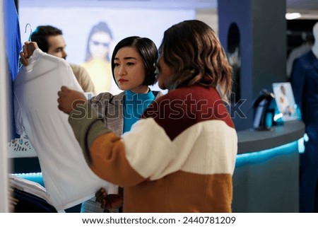 Diverse girlfriends shopping in clothing store, holding shirt on hanger and examining style. Two women friends choosing fashionable formal wear apparel in retail center boutique Royalty-Free Stock Photo #2440781209