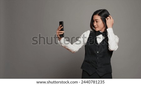 Hotel concierge taking photos on smartphone app in studio, having fun with pictures and working as a receptionist. Asian front desk staff using mobile phone to capture silly selfies. Camera A.