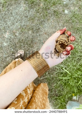 Henna hands with traditional Pakistani jewellery, including golden brown metal earrings called “jhumka” and copper brown bangles with matching brown dress  , and green grass background.