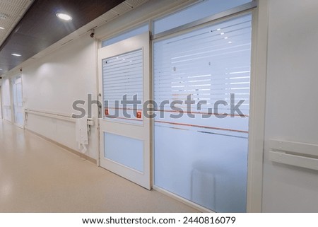 a bright hospital corridor with numbered doors, frosted glass windows, handrails, and a paper towel dispenser.