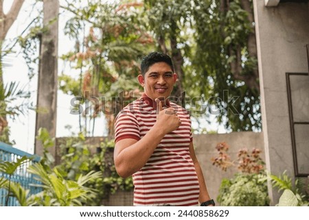 A jovial Asian man, dressed casually, stands outdoors with foliage in the background, giving a thumbs-up sign.