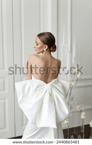 Photos featuring a slender, European-looking woman in a stylish white gown, her back elegantly bare, highlighting her poise and elegance in the pristine setting of a photo studio with white doors.
