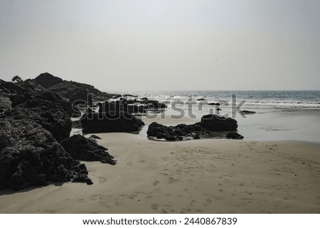 black or dark grey rocks at sea shores.  Beautiful view of a sandy beach with foot marks on a sunny day. Crab rocks at sea shore. Beach with rocks in low tide. Waves in Arabian sea in Goa, India.  Royalty-Free Stock Photo #2440867839