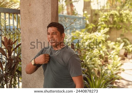 A middle-aged Asian man experiences discomfort from the extreme heat, attempting to cool down by fanning his shirt outside. Royalty-Free Stock Photo #2440878273