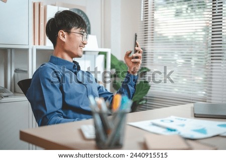 businessperson, document, job, manager, occupation, working, student, smart phone, corporate, freelance. A man is sitting at a desk with a cell phone in his hand. He is smiling and he is happy.