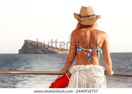 Blonde woman wearing a bikini and a hat, looking at an island while sailing on a boat in the Mediterranean Sea in summer