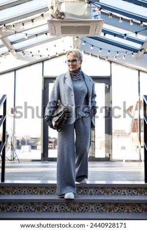 Attractive and stylish mature woman in grey pant suit with jacket and turtleneck, accessorized with handbag and white shoes. The perfect look for successful women over 50. Royalty-Free Stock Photo #2440928171
