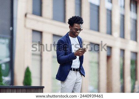 Stylish African American man in smart casual clothes, with mobile phone against backdrop of city. Fashionable guy with smartphone outdoors. Royalty-Free Stock Photo #2440939461
