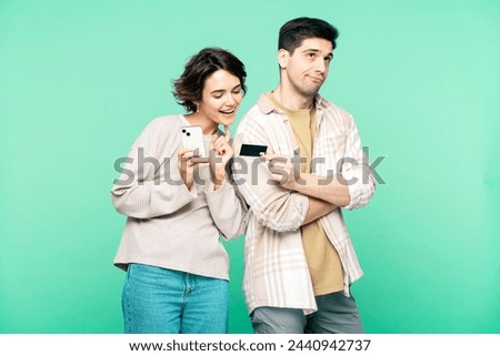 Funny married couple; woman with mobile phone reading off bank card number for shopping, while frustrated man giving credit card to his delighted wife, isolated on a turquoise background, looking away Royalty-Free Stock Photo #2440942737
