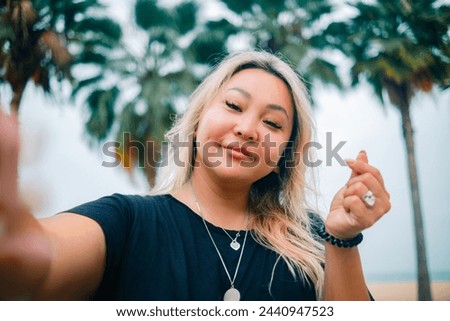 Young asian pretty woman shows heart sign making selfie with happy smile with ocean beach palms on background. High quality photo