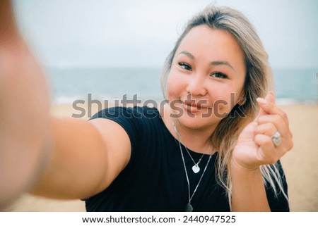 Young asian pretty woman shows heart sign making selfie with happy smile with ocean beach on background. High quality photo