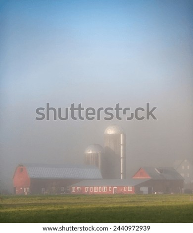 A red barn with silver silos in heavy fog on a farm in rural Vermont. Royalty-Free Stock Photo #2440972939