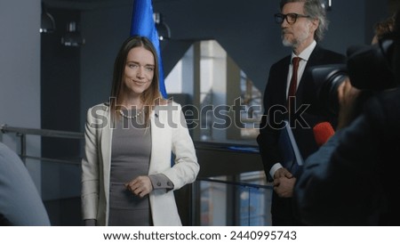 Positive female European politician smiles, poses for cameras and greets journalists. Media workers take pictures and broadcast on television. Representative of the European Union at press conference.