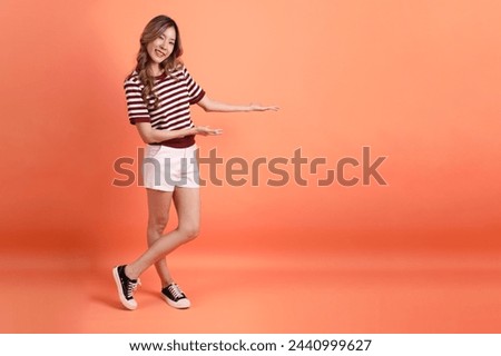 The young Asian woman in casual clothes with gesture of invitation or welcome on the orange background 