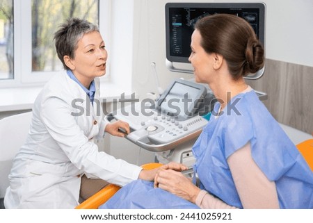 A doctor and patient converse during an ultrasound appointment, with the doctor operating the machine and the patient in a blue gown, seated comfortably.