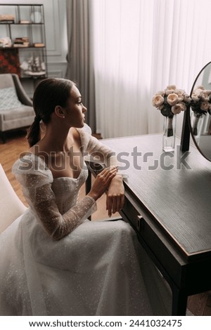 The person is wearing a wedding dress and the setting is indoors. The photo features a bride in bridal clothing with embellishments, sitting next to a wall.