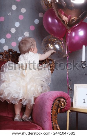 A beautiful one-year-old baby girl in a festive dress with a crown on her head is standing on a pink chair and touching a balloon. Back view. Vertical image. 