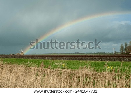The rainbow starts at the mill on a day with changeable weather. In the front, the train track near Adorp, The Netherlands. Typical Dutch picture.