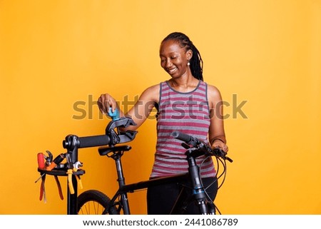 Energetic black woman making bike adjustments with tools and inspecting components. Sports-loving african american lady examining repair-stand for broken bicycle frame. Isolated yellow background. Royalty-Free Stock Photo #2441086789