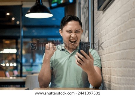 Asian man Engaging with Smartphone in a Modern Cafe Setting. Looking to good news on his phone concept