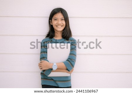 A young girl is holding a laptop in her arms and smiling. Concept of happiness and excitement, as the girl appears to be proud of her new device. The laptop itself is a symbol of modern technology