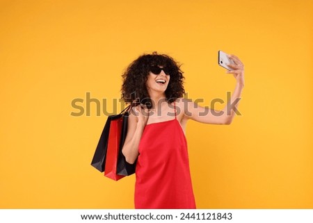 Happy young woman with shopping bags and stylish sunglasses taking selfie on yellow background