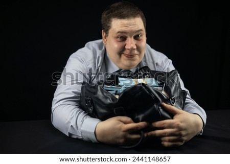 A male official hugged a bag full of dollars and looked happily at the camera on a black background. A joyful official with a black bag full of stacks of hundred dollar bills.