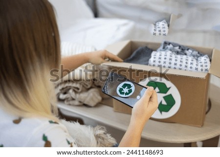 A woman holds a phone in her hands to take pictures of clothes folded in a box. A recycling sign is visible on the phone. Placing an advertisement on the Internet for the sale of old clothes.