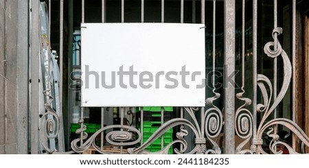 Square blank sign hanging on ornate wrought iron gate in front of a shop window. Suitable for business logos, opening hours, or welcome signs in a vintage setting
